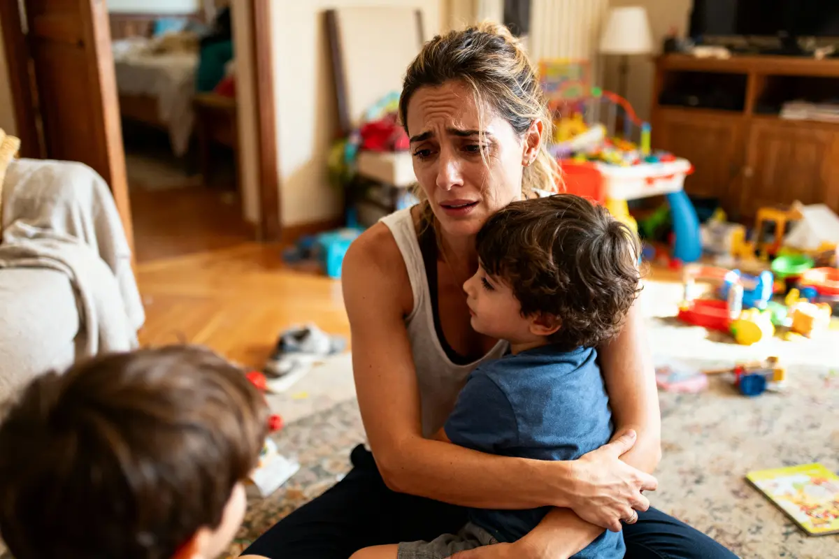 Une femme étreint deux enfants dans un salon en désordre, air préoccupé.