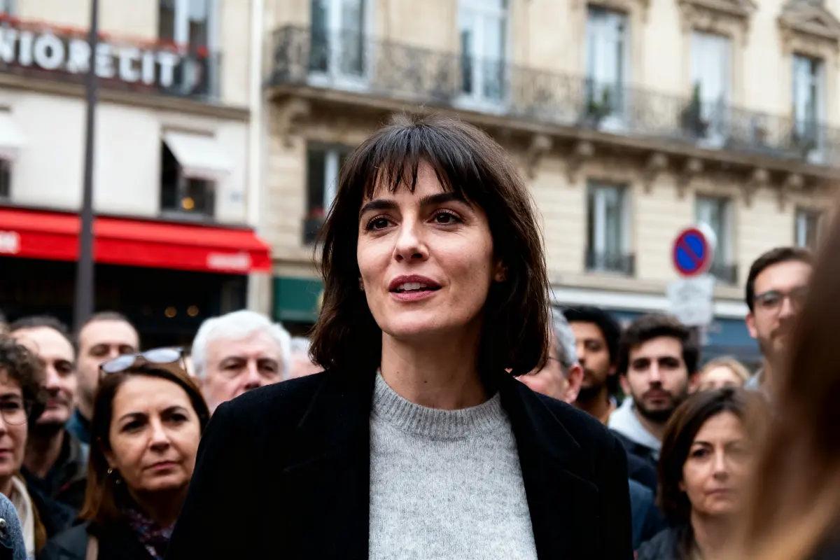 Groupe de personnes écoutant une femme souriante, en extérieur, devant bâtiments parisiens avec enseignes et balcon.