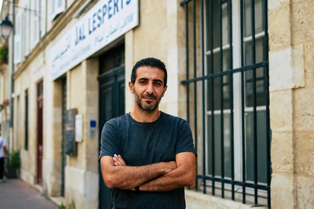 Homme debout devant bâtiment en pierre, bras croisés, expression neutre, fenêtre à barreaux, panneau illisible derrière.