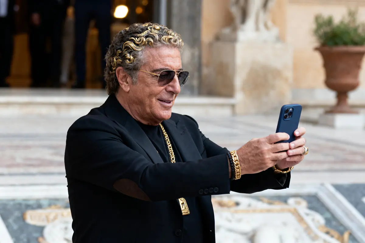 Homme en costume noir, cheveux bouclés, lunettes de soleil, prend photo avec smartphone, bracelets et collier dorés.
