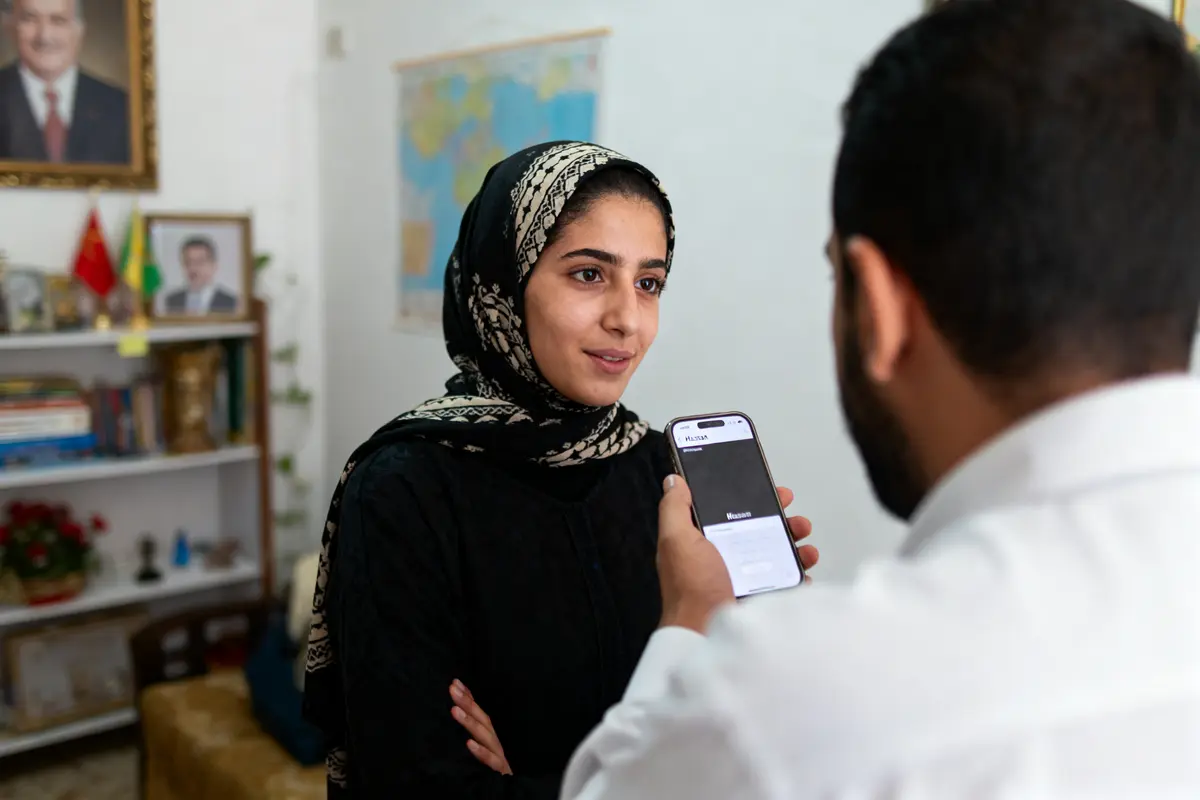 Un homme filme une femme en hijab avec un smartphone, dans une pièce décorée de livres et portraits.