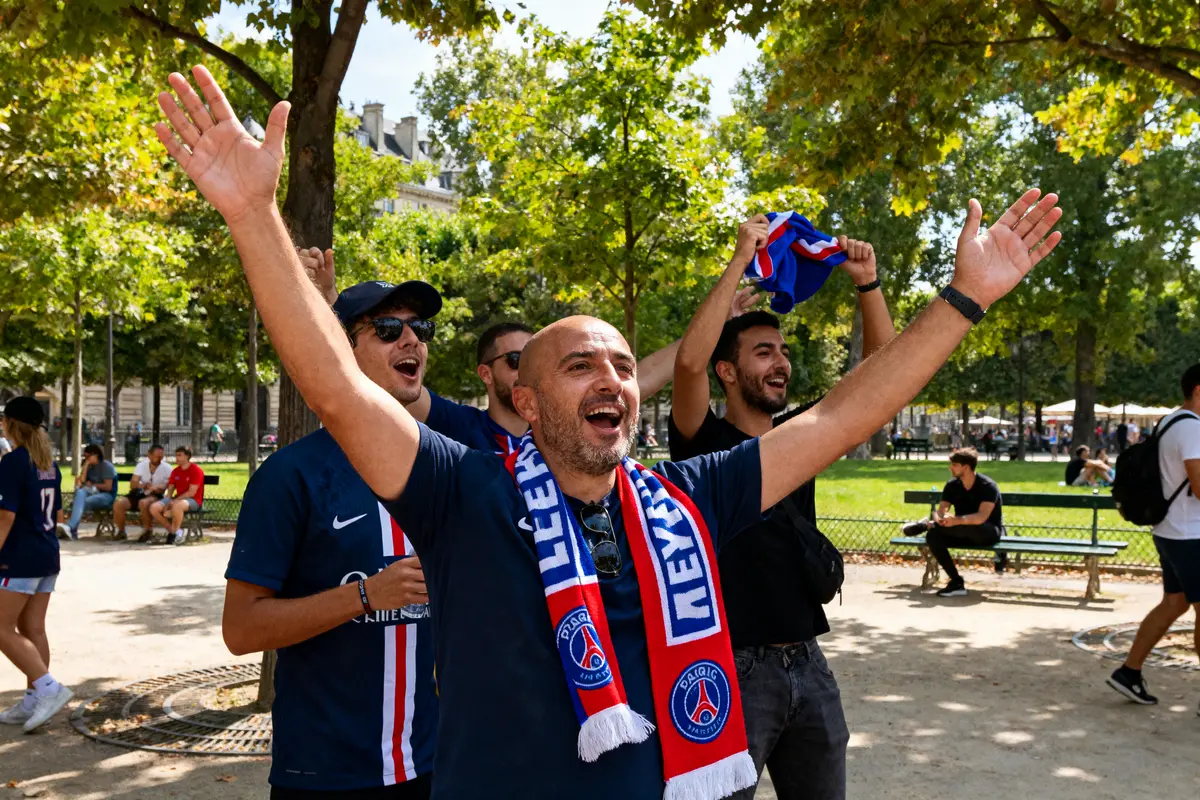 Supporters du PSG célébrant dans un parc, portant écharpes, souriants, bras levés, arbres en arrière-plan.