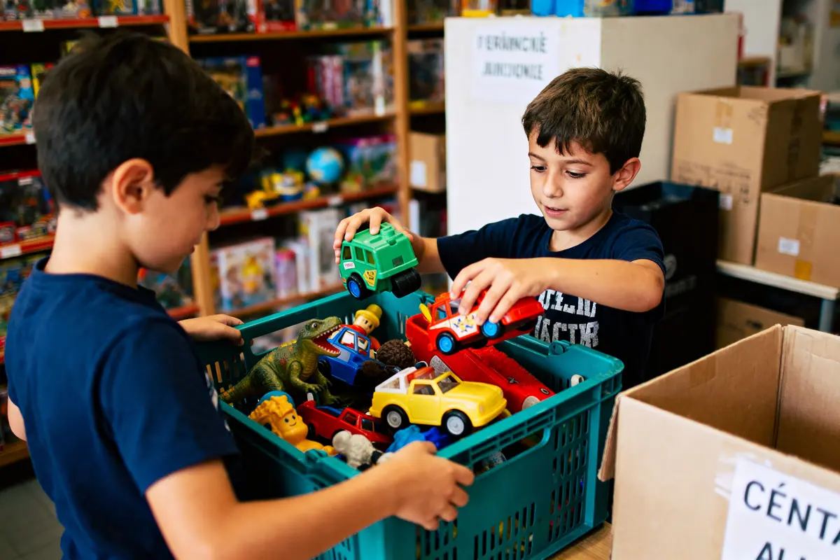 Deux enfants sélectionnent des jouets colorés dans une caisse en plastique, entourés de cartons et étagères.