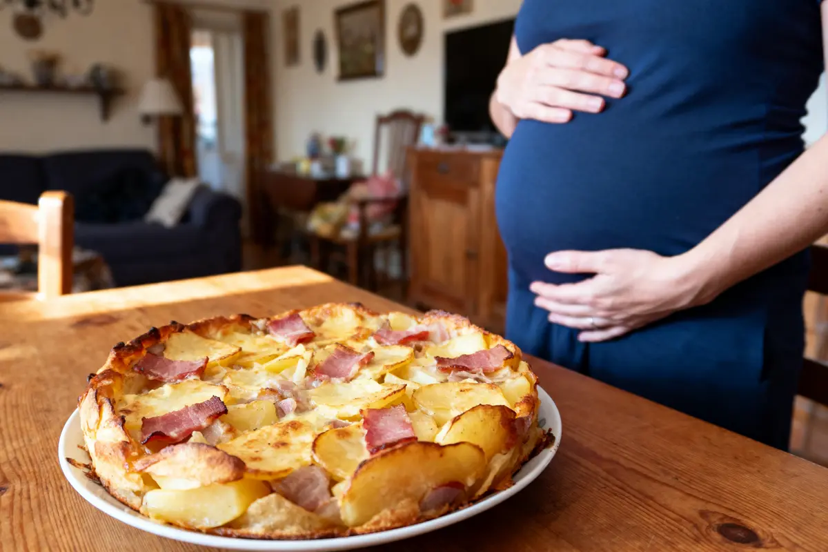 Plat de gratin avec du bacon sur table, personne enceinte touchant ventre en arrière-plan dans un salon.