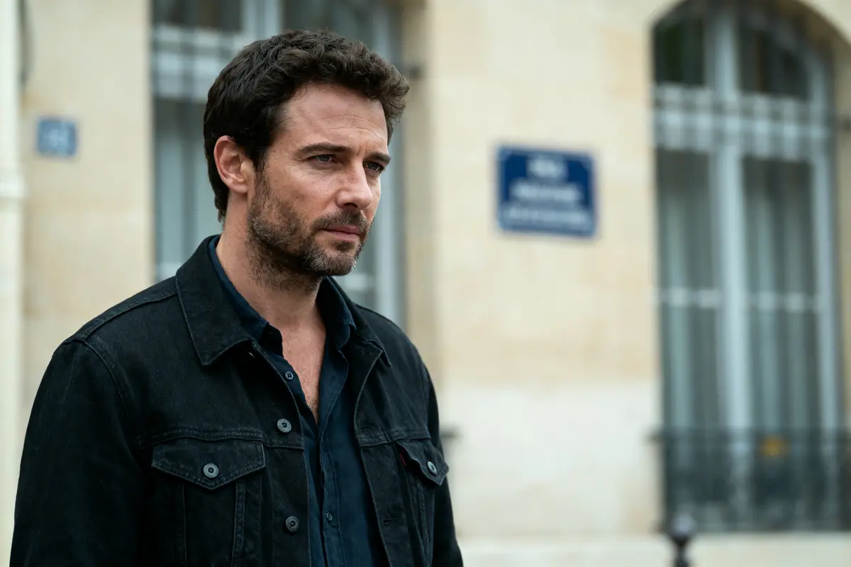 Homme barbu en veste noire devant bâtiment avec fenêtres et plaque de rue floue en arrière-plan.