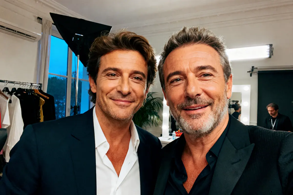 Deux hommes en costumes sourient, posant pour un selfie dans une loge bien éclairée.