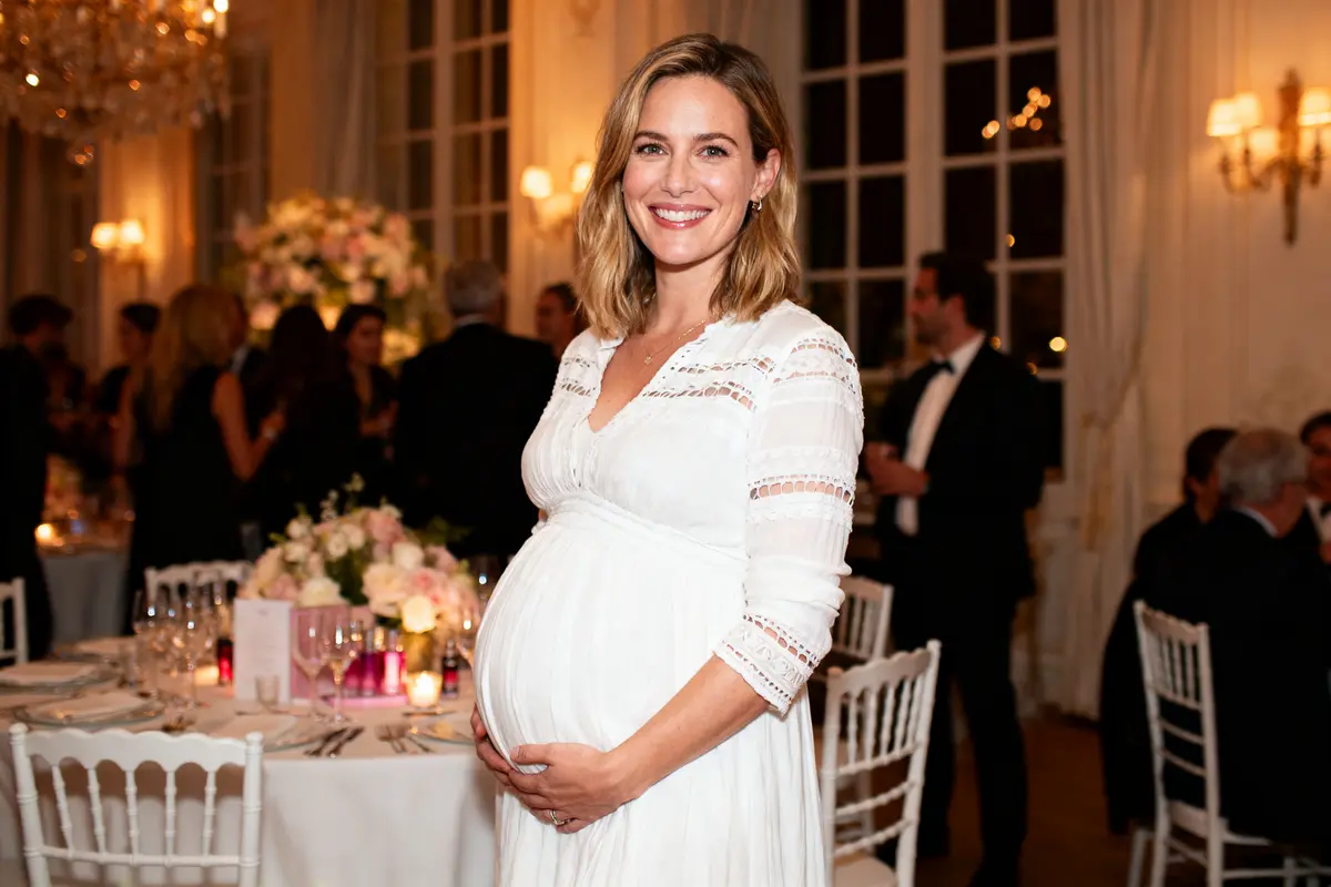 Une femme enceinte souriante en robe blanche dans une élégante réception avec invités et table décorée.