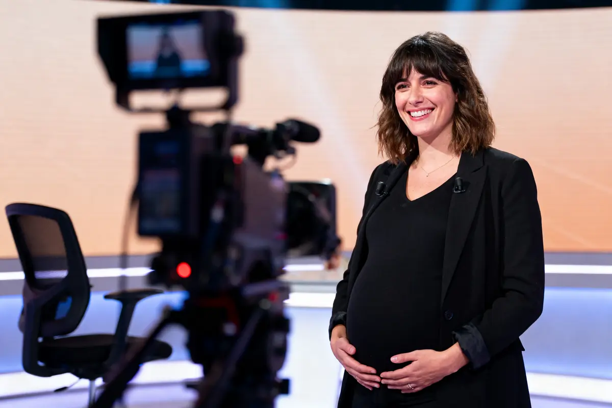 Femme enceinte souriant devant une caméra dans un studio de télévision, portant une tenue noire.