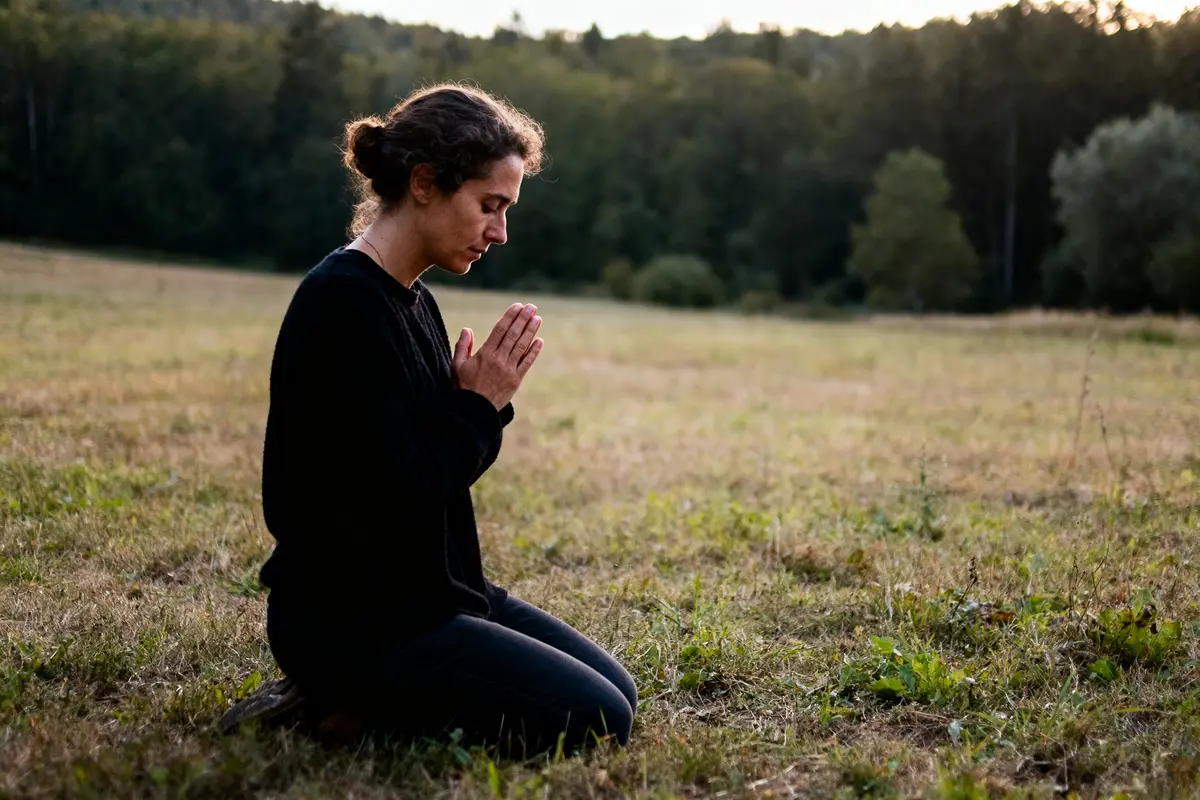 Personne méditant dans un champ, les mains jointes, forêt en arrière-plan, ambiance paisible et naturelle.