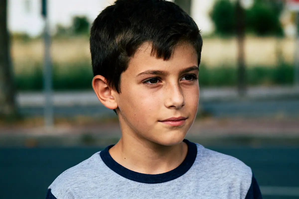 Jeune garçon aux cheveux bruns, portant un t-shirt gris, regardant au loin, arrière-plan flou.