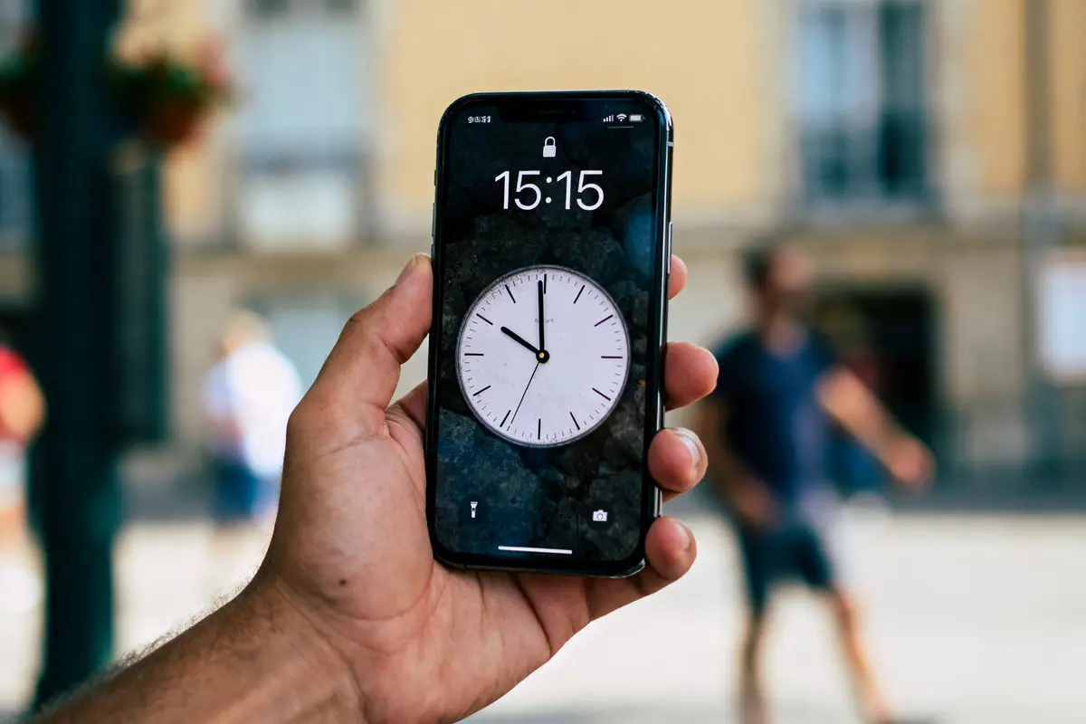 Main tenant un smartphone affichant l'heure 15:15, fond urbain flou, écran verrouillé avec cadran d'horloge.